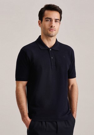 Junger Mann, der ein schwarzes kurzärmeliges Poloshirt und schwarze Hose trägt, steht vor einem einfarbigen hellen Hintergrund mit den Händen in den Taschen.