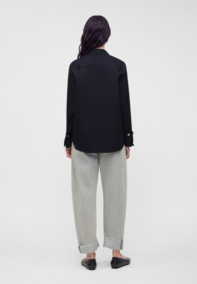 Femme aux longs cheveux foncés portant un t-shirt noir à manches longues, un jean gris clair retroussé et des chaussures plates noires, debout, de dos.