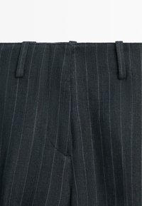 Zwarte pantalon met fijne krijtstrepen, voorzien van lussen voor een riem en een verborgen ritssluiting aan de voorkant, weergegeven vanaf de taille omhoog.