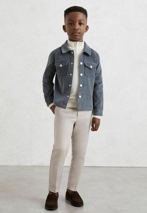 Poiss seisab heledal taustal, kandes halli nööbitavat jopet, beeže pükse, pruune kingi ja valget lukuga kampsunit.