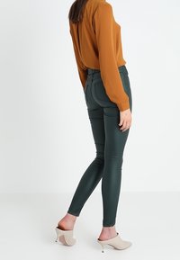Tummanvihreät slim-fit-housut, joissa on sileä pinta, yhdistettynä oranssiin puseroon ja vaaleanbeigeihin korkokenkiin, nähtynä takaa valkoista taustaa vasten.