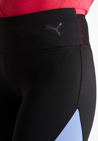 Leggings negras con una cinturilla ancha, que presentan un pequeño logo de Puma en gris y un panel azul claro en el lateral. Fabricadas en un material elástico.