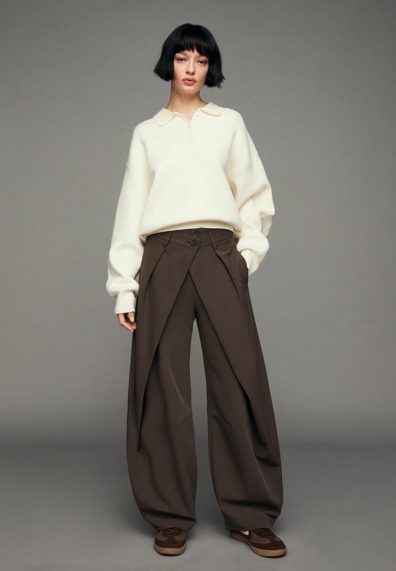 Krémový pletený svetr s límečkem, doplněný volnými tmavě hnědými kalhotami s jedinečným složeným designem a zapínáním na knoflíky. Hnědá obuv.