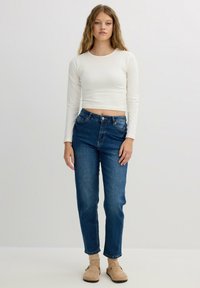 Top corto de manga larga blanco, jeans rectos de talle alto en azul y botas de tobillo beige. Diseño simple y casual.