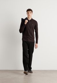 Mørkebrun langærmet polo skjorte med krave, med et lille logo, parret med sorte bukser og sko, båret af en stående mandlig model.