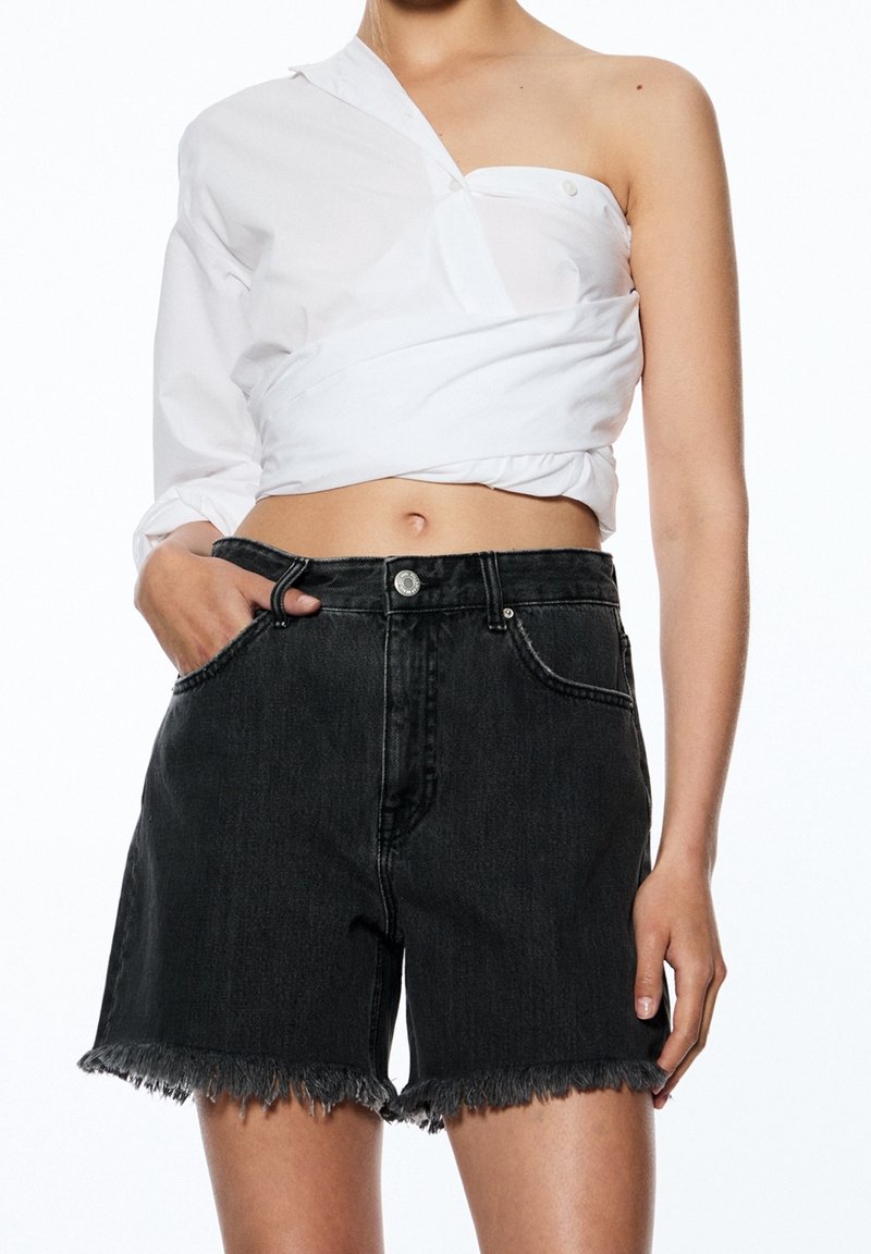 Femme portant un haut blanc asymétrique et un short en jean noir taille haute à ourlets effilochés, main dans la poche, montrée des épaules aux cuisses.