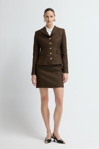 Bruine tweed jas met een brede revers, gouden knopen en bijpassende nauwsluitende rok. Zwarte slingback schoenen met een spitstuiten en gouden accenten.