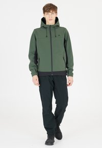 Grön huva mjukskaljacka med svarta detaljer, framdragkedja och två sidofickor. Matchat med svarta byxor och stövlar. Slät textur.