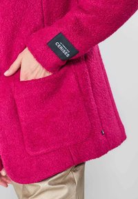 Fuchsia fleece jas met een patchzak, voorzien van een zwart label met witte tekst. Zachte textuur en comfortabele pasvorm. Gouden broek zichtbaar daaronder.