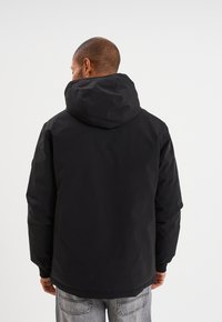 Chaqueta negra con capucha, con un exterior liso y resistente al agua, y puños elásticos, llevada sobre unos jeans de mezclilla gris claro. Vista trasera mostrada.