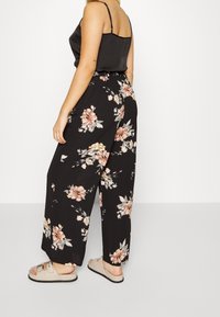 Pantalons larges à motifs noirs avec des designs floraux en rose et beige, associés à un haut sans manches noir et des sandales à glisser beiges.