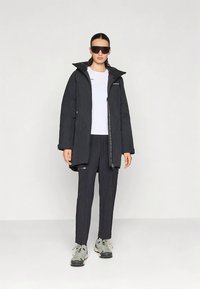 Γυναίκα που φοράει μαύρη κουκούλα παρκά, άσπρο πουκάμισο, μαύρο παντελόνι, γκρι αθλητικά παπούτσια και μεγάλα μαύρα γυαλιά ηλίου, που στέκεται μπροστά από ένα απλό ανοιχτό φόντο.