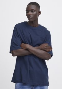 Tricou din bumbac bleu marin, cu mâneci scurte, guler rotund și un buzunar pe piept. Textură netedă, croială lejeră și detaliu subtil al brandului pe buzunar.