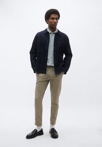 Σκούρο τζιν μπουφάν με δύο τσέπες στο στήθος πάνω από ανοιχτόχρωμο ριγέ πουκάμισο με κουμπιά, συνδυασμένο με μπεζ παντελόνι jogger και μαύρα μοκασίνια.