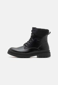 Bota de cuero negro hasta el tobillo con un collar de tela negra texturizada, punta redonda y cierre de cordones. Cuenta con una suela de goma resistente y una lengüeta.