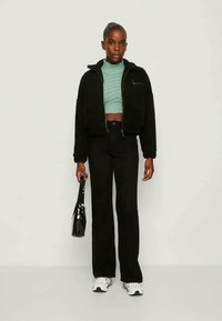 Black cropped jacket over a light blue knitted top, paired with black flared pants and white sneakers. The model holds a black handbag.