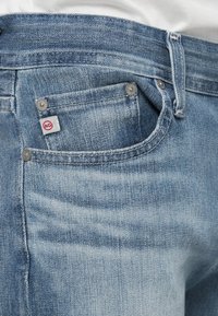 Gros plan sur la poche d’un jean denim bleu clair avec une petite étiquette de marque AG blanche et rouge et des rivets métalliques.