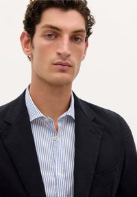 Blazer azul marino oscuro sobre una camisa de rayas azul y blanca, con un cuello puntiagudo y un cierre de botones, que cuenta con un detalle de bolsillo en el pecho.