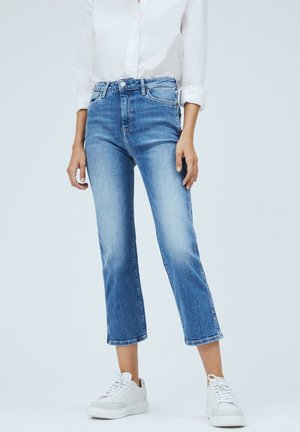 Vrouw draagt lichtblauwe cropped jeans, wit overhemd met knopen en opgerolde mouwen en witte sneakers, staand tegen een effen achtergrond.