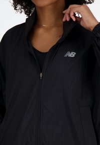 Veste légère noire en tissu texturé. Caractéristiques : fermeture éclair complète et logo New Balance en gris sur la poitrine.