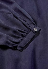 Marineblauer Stoff mit strukturiertem Finish, ausgestattet mit gerafftem Ärmel-Detail und einem einzelnen Knopfverschluss am Bündchen. Weiches, fließendes Material.