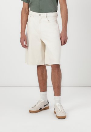 Shorts en denim blanc avec une coupe décontractée, présentant une fermeture à bouton à l'avant, des passants de ceinture et des poches latérales. Assortis avec des baskets crème et noires.