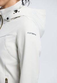 Hvid softshell-jakke med hætte, der har et buet sømdesign, lynlås-lukning og sort logo på ærmet. Letvægts og glat tekstur.