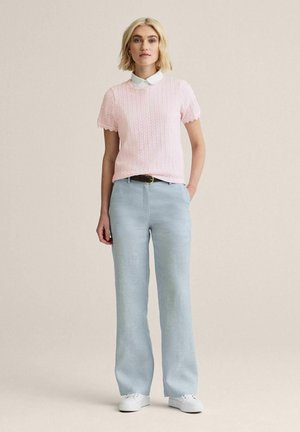 Kvinde står med hånden i lommen, iført lyserød kortærmet striktrøje over hvid skjorte med krave, lyseblå bukser og hvide sneakers.