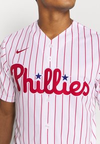 Biała koszulka baseballowa w czerwone paski. Napis "Phillies" dużymi czerwonymi literami z niebieskimi gwiazdkami. Logo Nike na lewej piersi, trzy białe guziki.