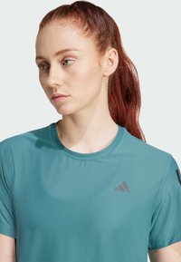 Tirkizna sportska majica kratkih rukava, okruglog izreza i teksturirane tkanine. Sadrži mali crni logotip na lijevoj strani prsnog područja.