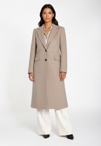 Beige kappa i ullblandning med en skräddarsydd design, spetsig krage, knäppning med en knapp och två framfickor, kombinerad med vida vita byxor.