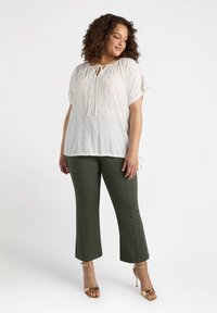 Blusa bianca con scollo a cravatta, maniche corte e dettagli oro; abbinata a pantaloni verdi oliva a trama corta e sandali con tacco oro.