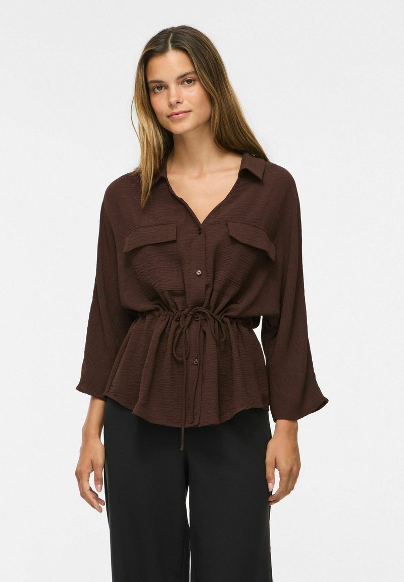 Femme portant un chemisier ample marron foncé boutonné avec une taille cintrée et un pantalon noir, debout devant un fond clair uni.