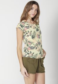 Bluzka z krótkim rękawem w jasnym żółtym kolorze w kwiatowy wzór z zielonymi liśćmi i różowymi kwiatami, zestawiona z oliwkowymi szortami z dwiema przednimi kieszeniami.