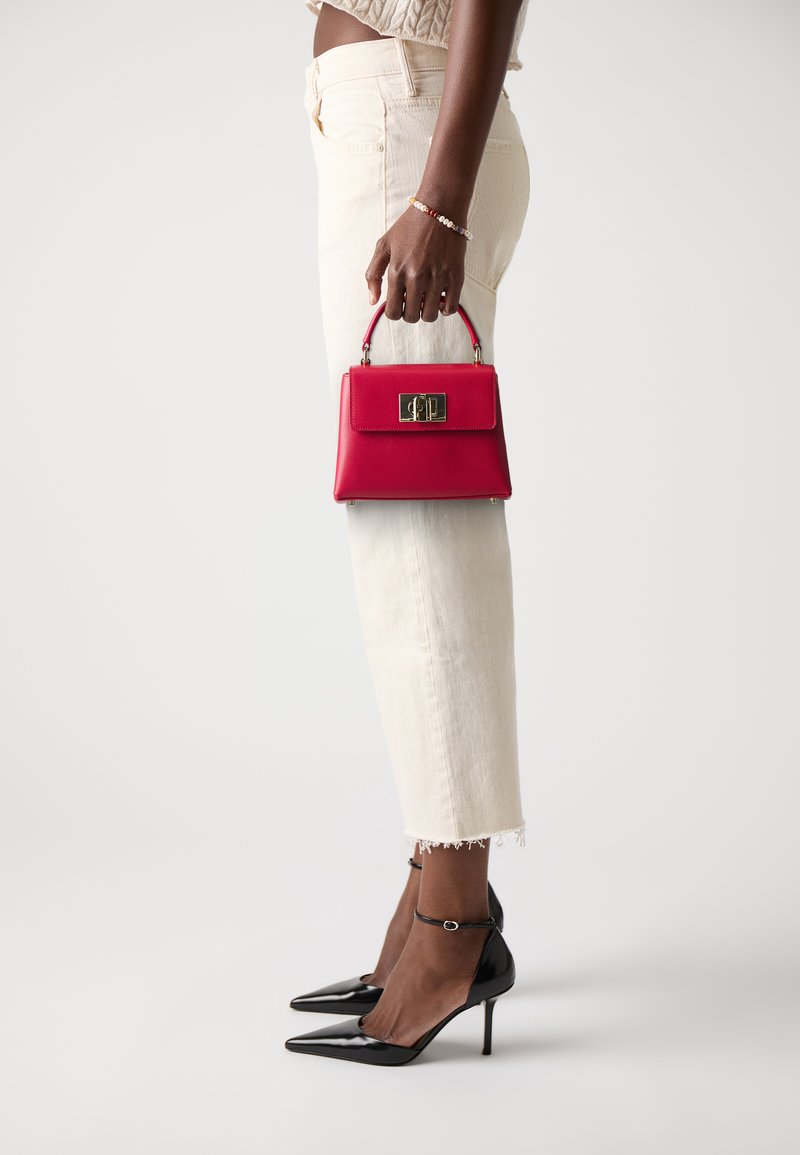 Sac en cuir rouge avec un fermoir doré, tenu par une personne portant un jean court crème et des escarpins noirs à bout pointu. Texture lisse, design structuré.