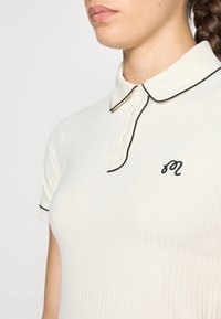 Bílá žebrovaná polokošile s černým lemovaným límcem a knoflíky, s malým černým vyšitým logem na hrudi. Krátké rukávy.