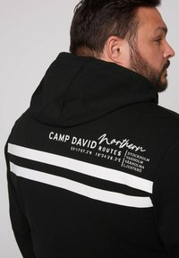 Sudadera negra con estampados gráficos en blanco en la parte posterior, que incluye texto y coordenadas, junto con dos franjas horizontales blancas.