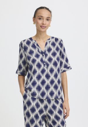 Donna che indossa un top a maniche corte e pantaloni larghi con motivo coordinato blu e bianco, in piedi con una mano in tasca.