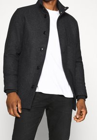 Uomo che indossa un cappotto grigio scuro con bottoni sopra una camicia bianca e pantaloni neri, in piedi contro uno sfondo chiaro e neutro.