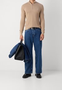 Uomo che indossa un maglione beige con zip, jeans blu, scarpe nere, che tiene in mano una giacca di denim e una borsa di pelle nera in un ambiente da studio.
