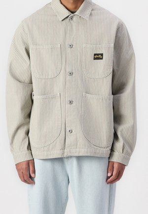 Man wearing beige and white vertical striped button-up jacket with four pockets, paired with light blue jeans.