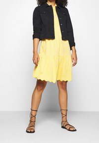 Sort denimjakke med to forlommer, lagdelt over en gul blomsterkjole med flere lag i skørtet og bølget kant, kombineret med sorte sandaler med remme.