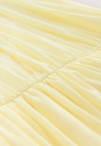 Pehme keltainen kangas, jossa on kerättyjä, rypytettyjä ompeleita, jotka luovat vaakasuoria taitoksia ja tekstuuria.
