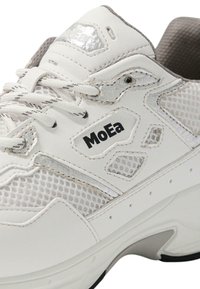 Weißer Sportschuh mit Mesh-Einsätzen, silbernen Akzenten und einer dicken Sohle. Die Marke "MoEa" ist an der Seite sichtbar. Strukturierte Details sind vorhanden.