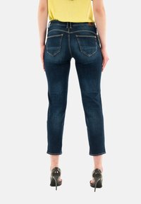Jeans de mezclilla azul oscuro con corte slim, que cuentan con dos bolsillos traseros, costuras visibles y un largo recortado. Usados con zapatos de tacón alto.
