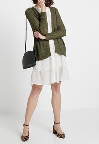 Cardigan testurizzato verde oliva sopra un vestito bianco con orlo a volant, abbinato a tacchi con stampa leopardata e una piccola borsa a tracolla nera.