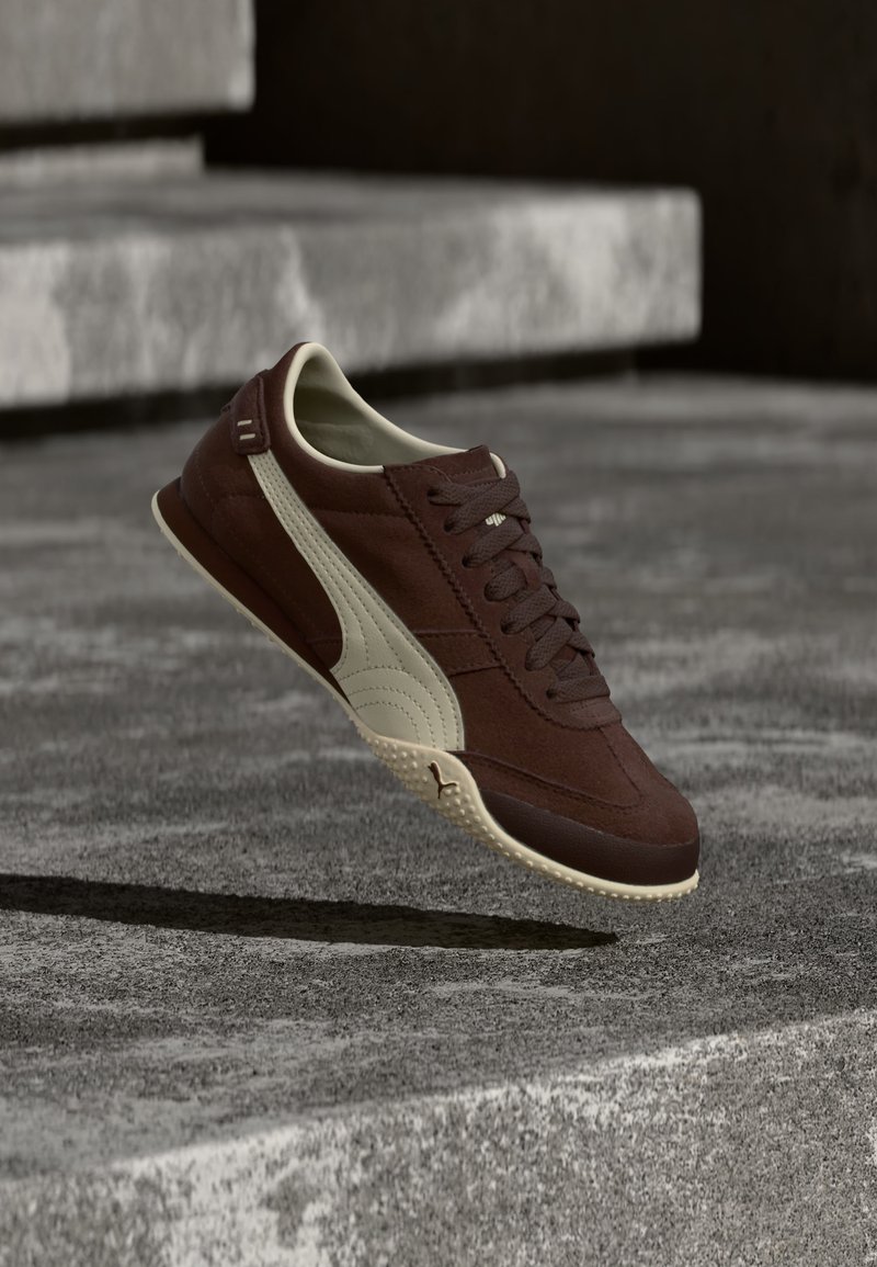 Καφέ αθλητικό παπούτσι με μπεζ σόλα και πλευρική ρίγα που αιωρείται πάνω από σκυρόδετα σκαλοπάτια σε φυσικό φως.