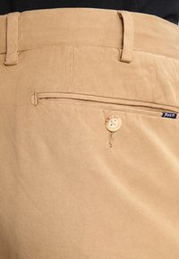 Khakifärgade byxor i bomull med en jämn textur. Har en bakficka med knappstängning och en liten märketsdetalj.