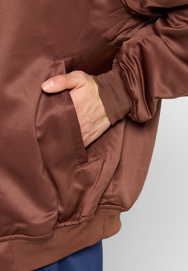 Giacca di raso marrone con una texture liscia, dotata di una tasca laterale e polsini a costine. L'orlo è elasticizzato per una vestibilità perfetta.