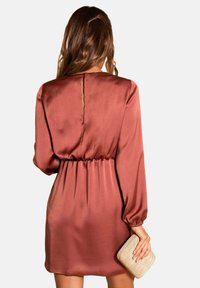 Robe en tissu lisse de couleur rouille, avec de longues manches, une taille froncée et un dos en forme de serrure. Un sac à main en osier est tenu dans une main.